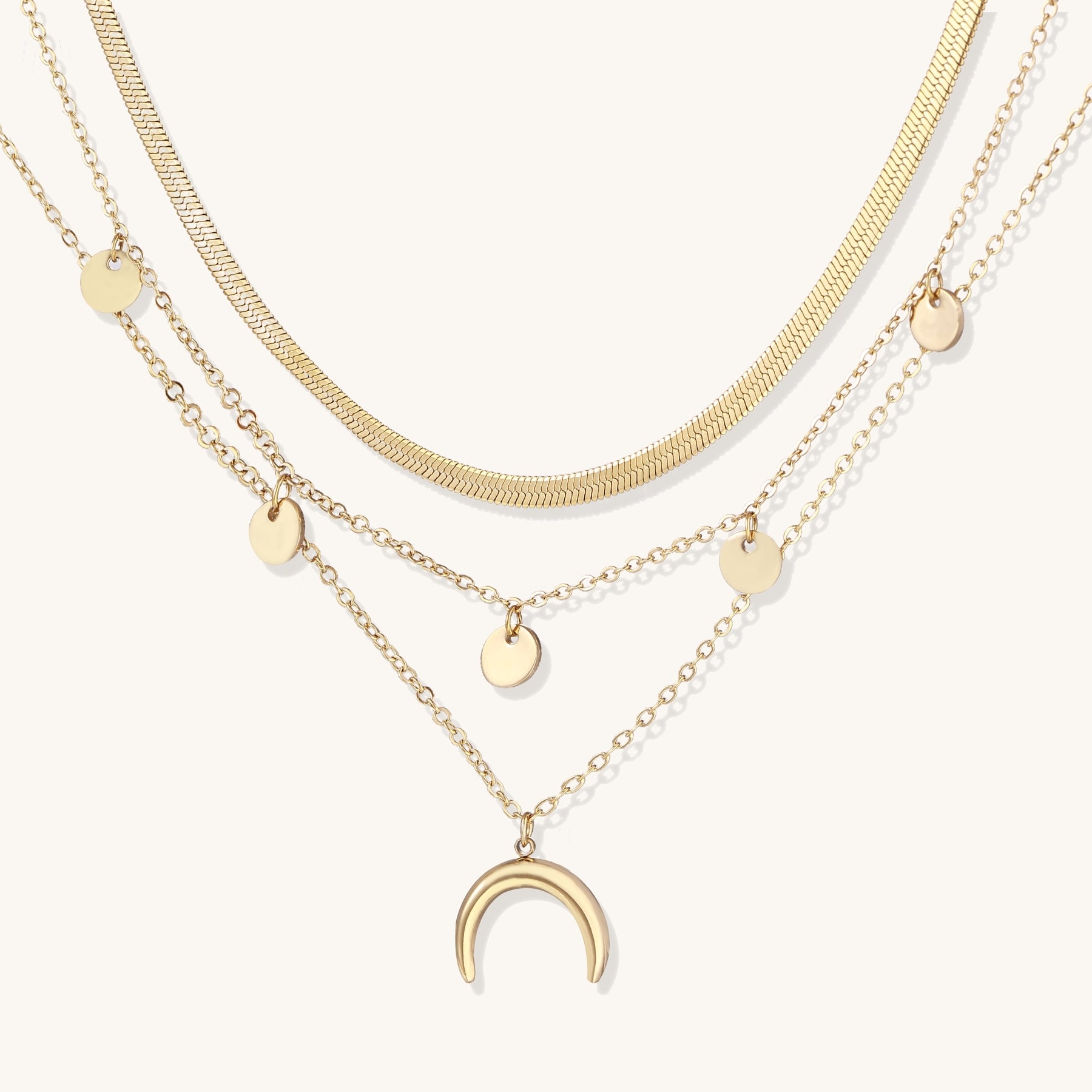 Gold-plated layered necklace set featuring herringbone chain, disc pendants, and crescent moon charm on beige background.