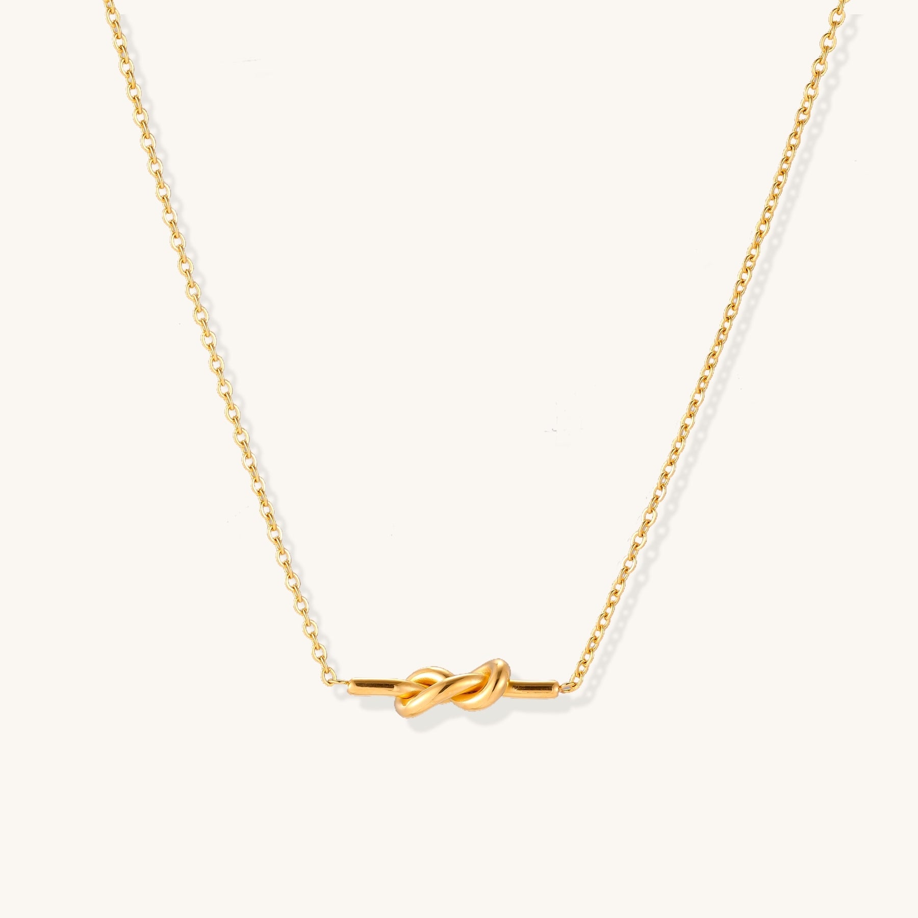Dainty 18K gold plated stainless steel chain necklace with minimalist knot pendant on cream background.